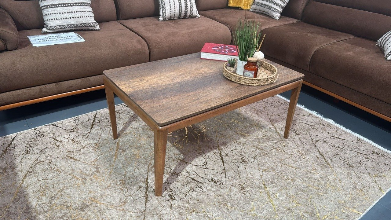 Cleo Coffee Table with a wooden finish and tapered legs, placed on a patterned rug in front of a brown sectional sofa.