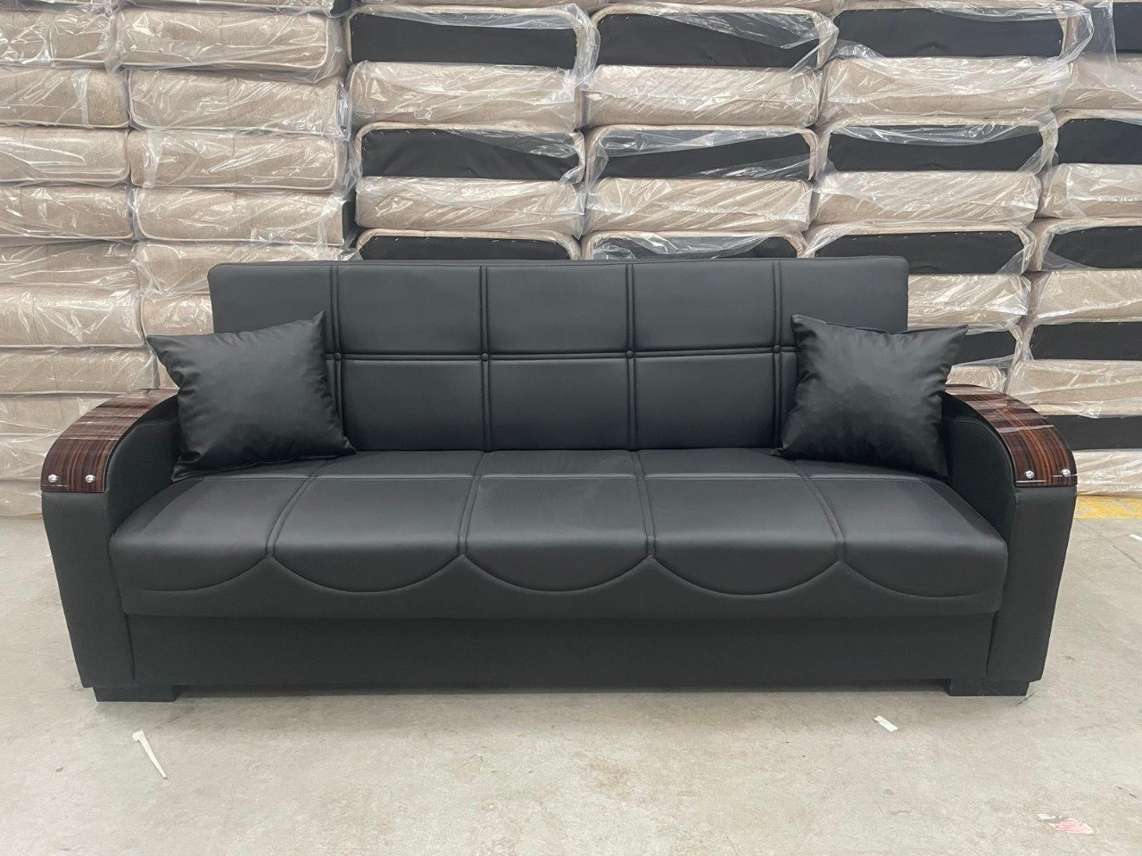 Black leather sofa with wood-accented armrests and two pillows, set against a backdrop of stacked, plastic-wrapped couches.