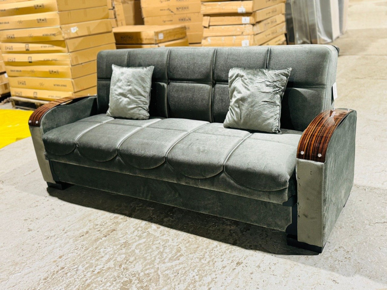 A grey sofa with wooden armrests and two pillows in a warehouse.