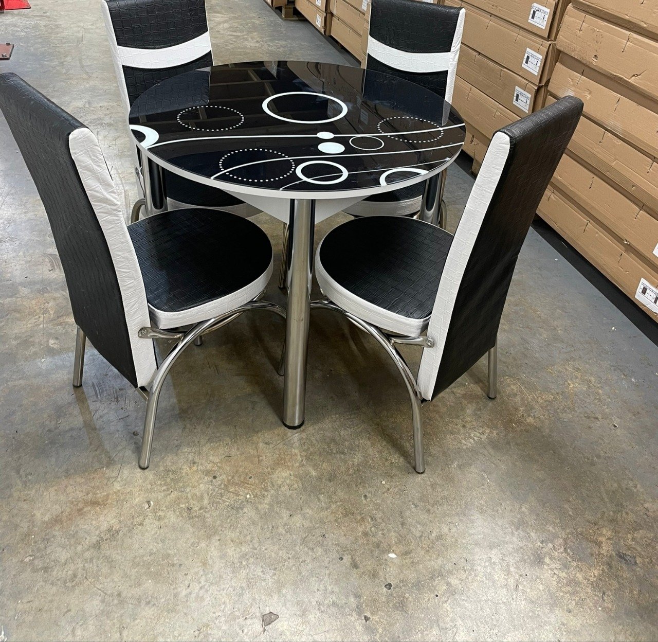Round glass table with black and white circle pattern and four black-and-white cushioned chairs in a warehouse setting.