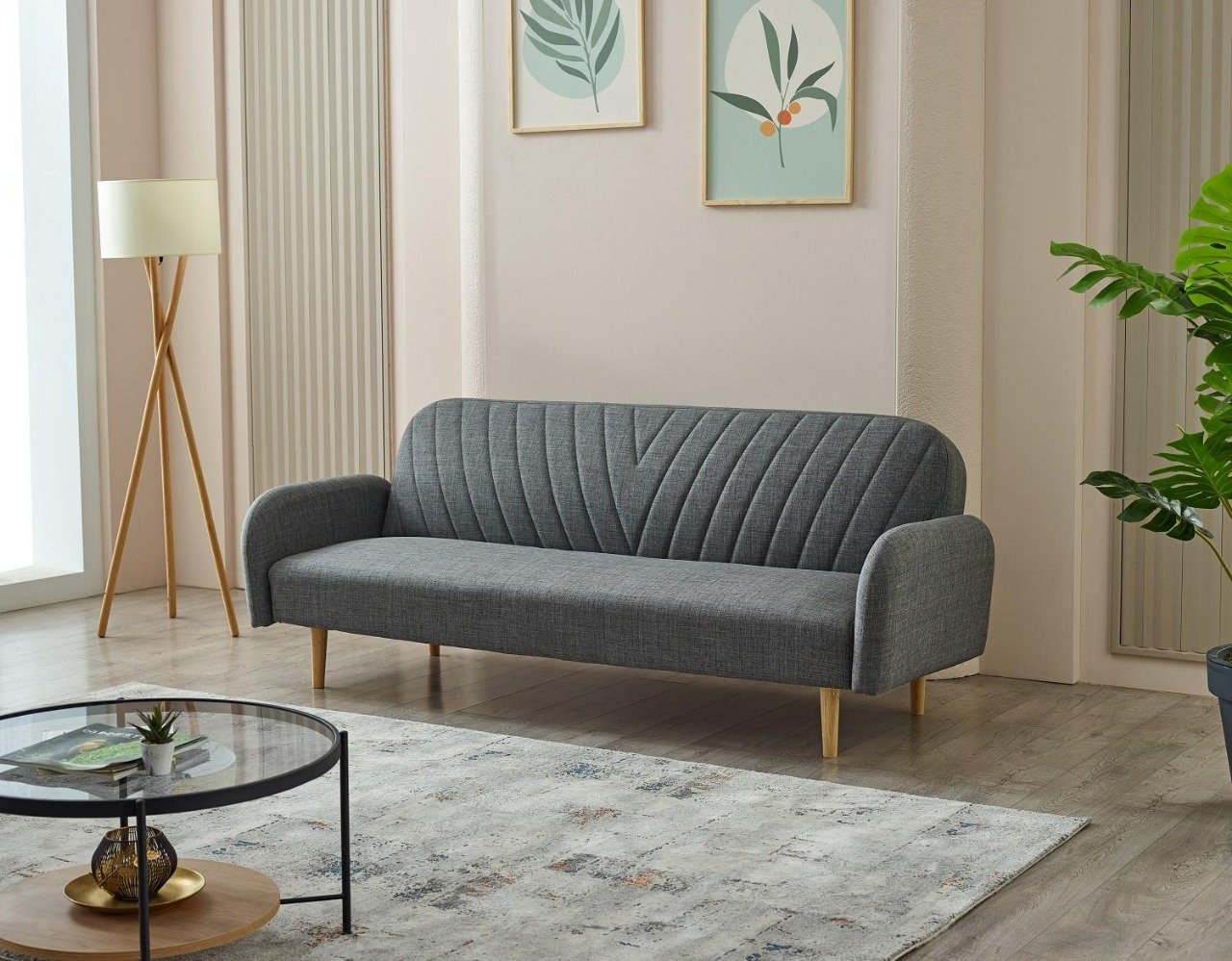 A modern living room with a gray sofa, tripod lamp, botanical artwork, round glass coffee table, and a large green plant.